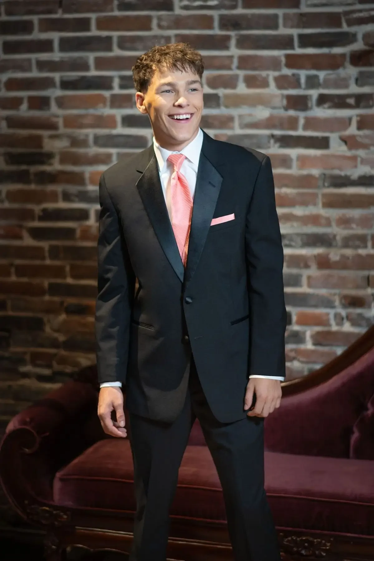 A young man in a formal suit with a pink tie and pocket square stands smiling in front of a brick wall, exuding confidence and joy.