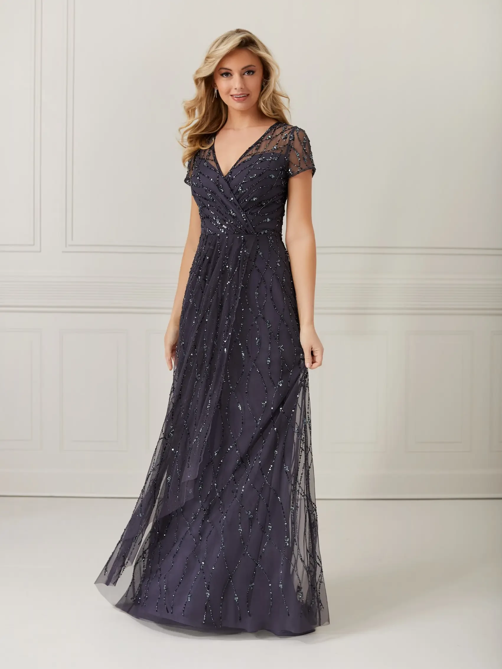 A woman in an elegant, embellished navy gown with short sleeves and a V-neckline stands in a bright room, exuding grace and confidence.