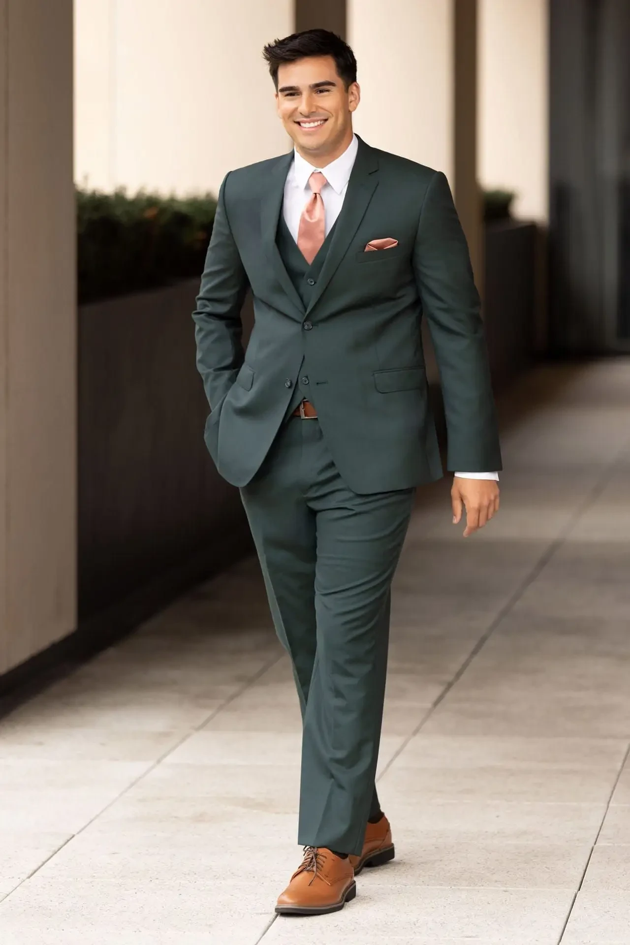 A man in a dark green suit with a matching vest and pink tie walks confidently along a modern outdoor corridor. His brown shoes complement the outfit.