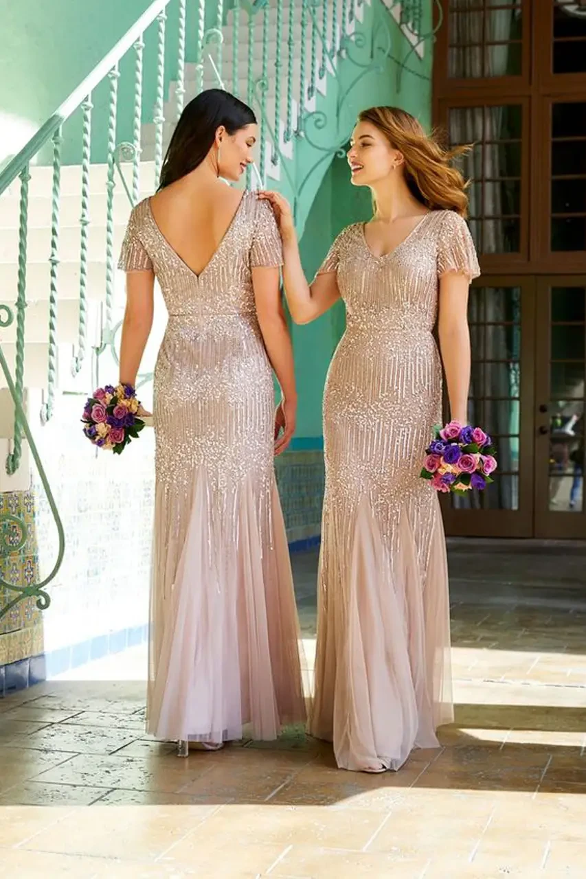 Two women in shimmering, floor-length gowns stand on a sunlit terrace, holding vibrant bouquets. They appear joyful, engaged in conversation.