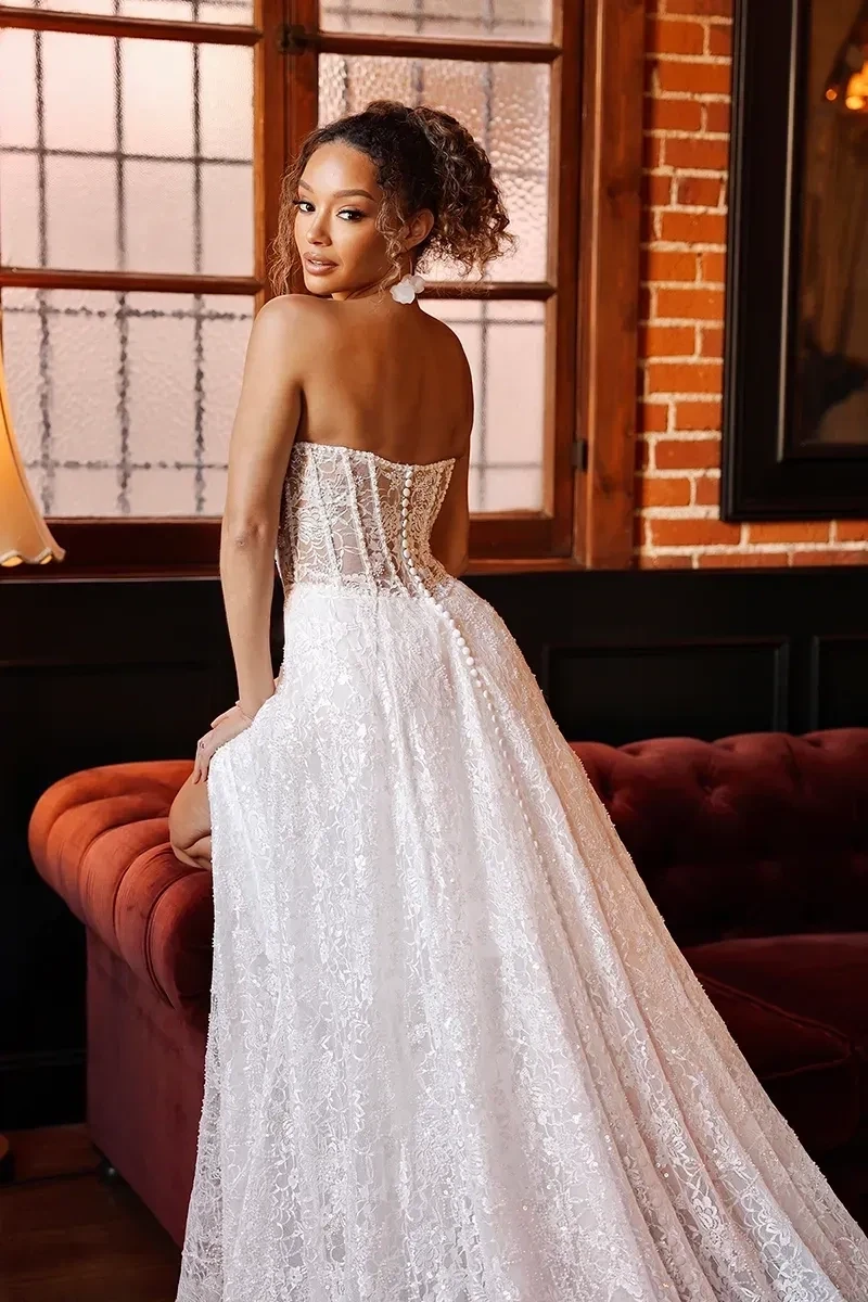 Model is wearing a strapless sheath gown with allover sparkle lace, a corset-style bodice, an A-line skirt, and a high leg slit.