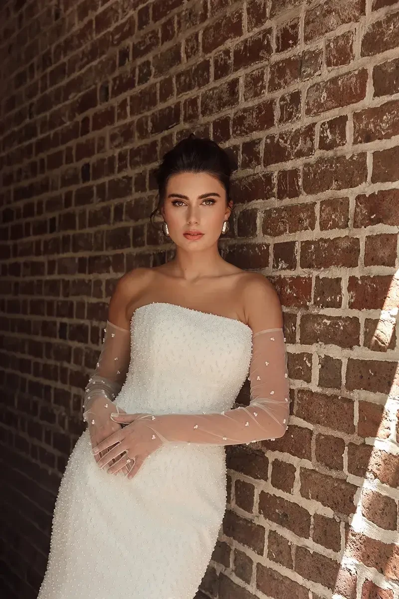 A woman in an elegant, strapless white dress with sheer sleeves leans against a sunlit brick wall, exuding confidence and poise.