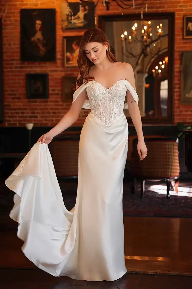 A woman wearing an elegant off-shoulder wedding dress with a fitted bodice and a flowing train, standing indoors in a stylishly decorated room.