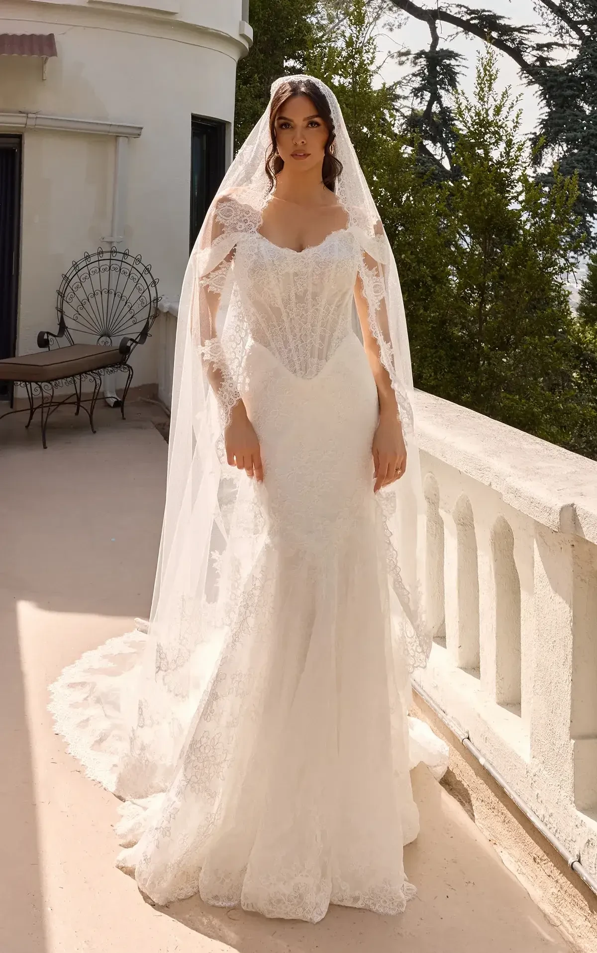 A woman in a white wedding dress with a long veil stands on a balcony, framed by greenery.