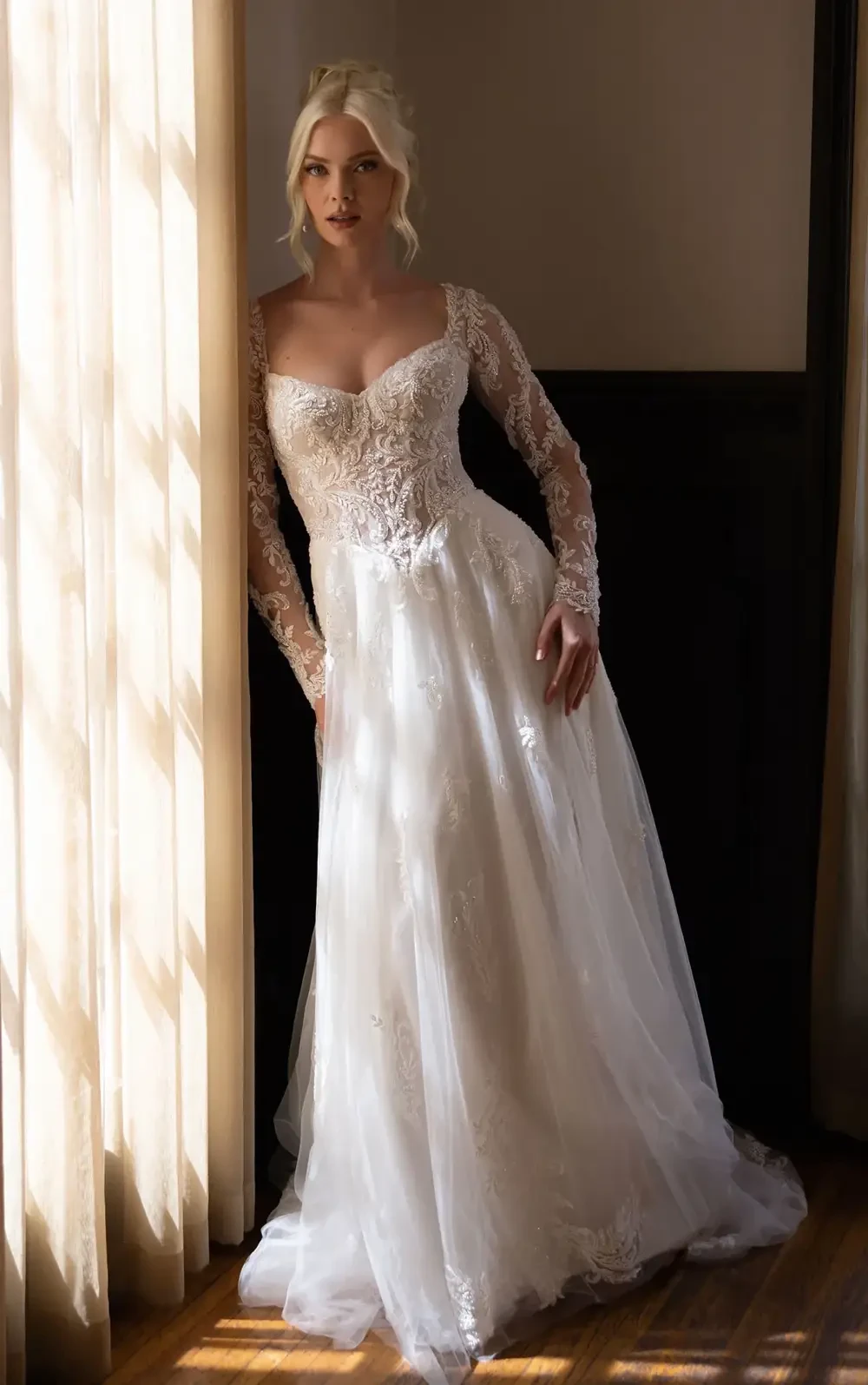 A model wearing a long-sleeved, lace wedding dress stands next to a window with soft natural light illuminating the scene.