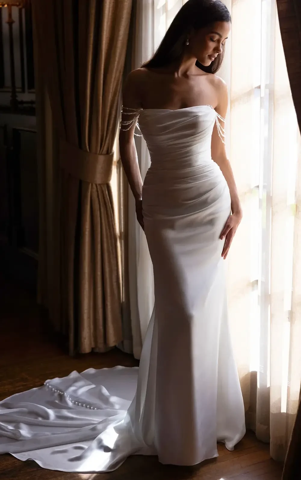 A woman in an elegant white dress stands by a window, with flowing fabric and a fitted silhouette, showcasing a glamorous style.