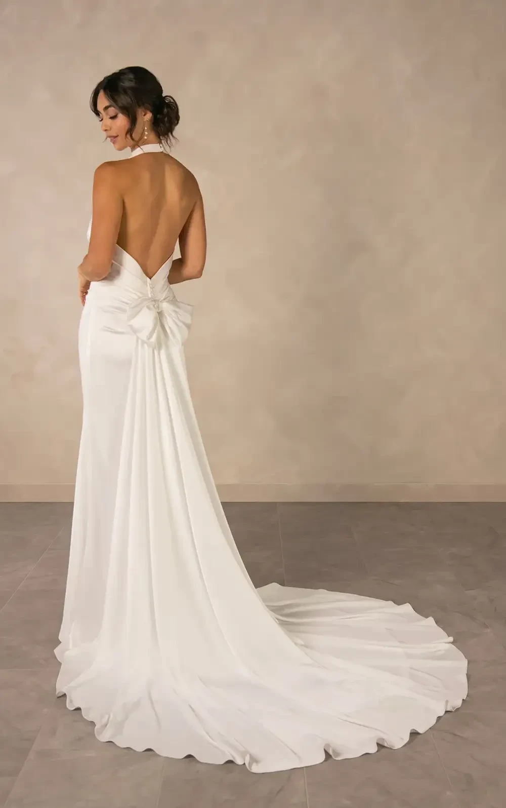 A woman in a backless, elegant white gown with a train and a bow at the waist, standing against a neutral background.
