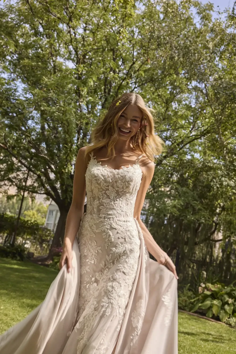 A smiling woman wearing a beautiful wedding dress stands in a garden, with sunlight filtering through the trees.