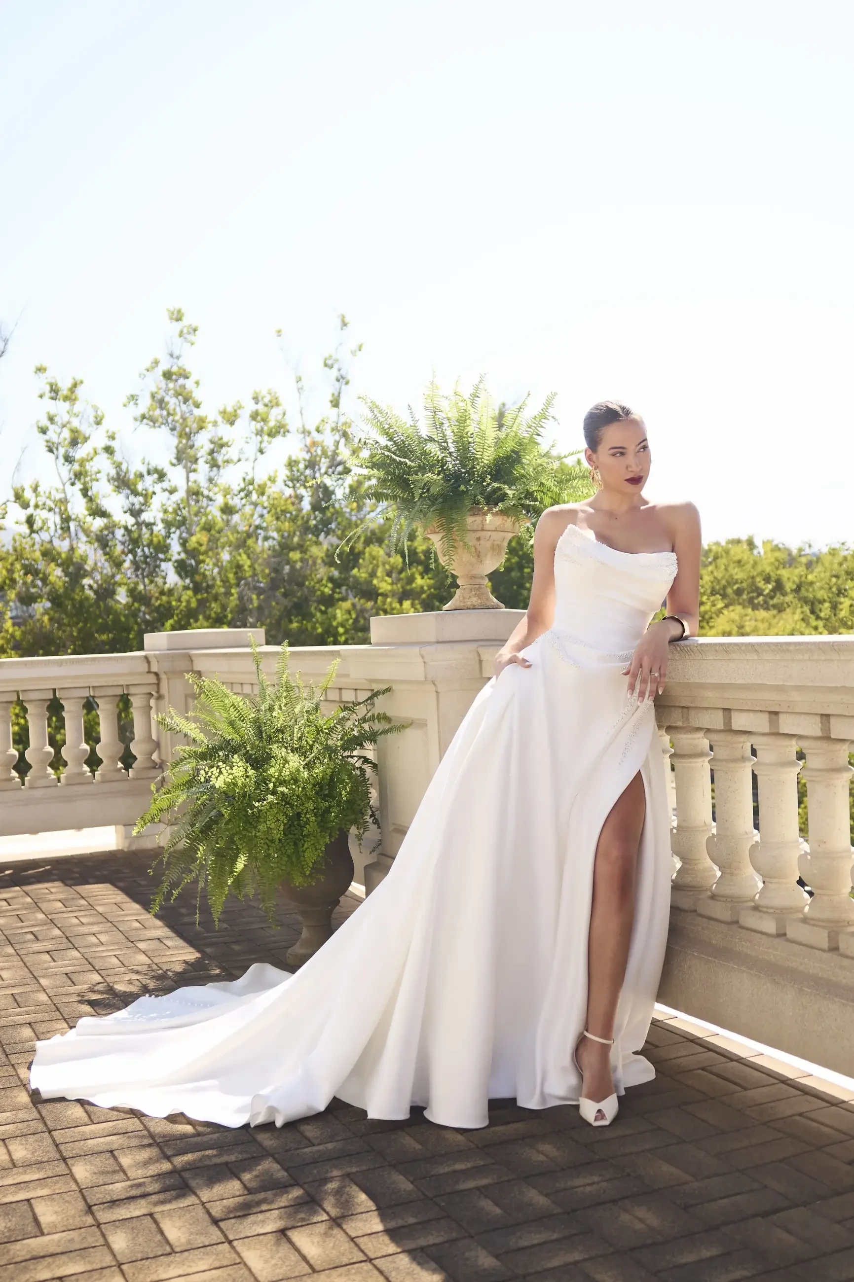 A woman in a white dress with a high slit stands on a balcony surrounded by greenery. The sunlight illuminates her as she poses elegantly.