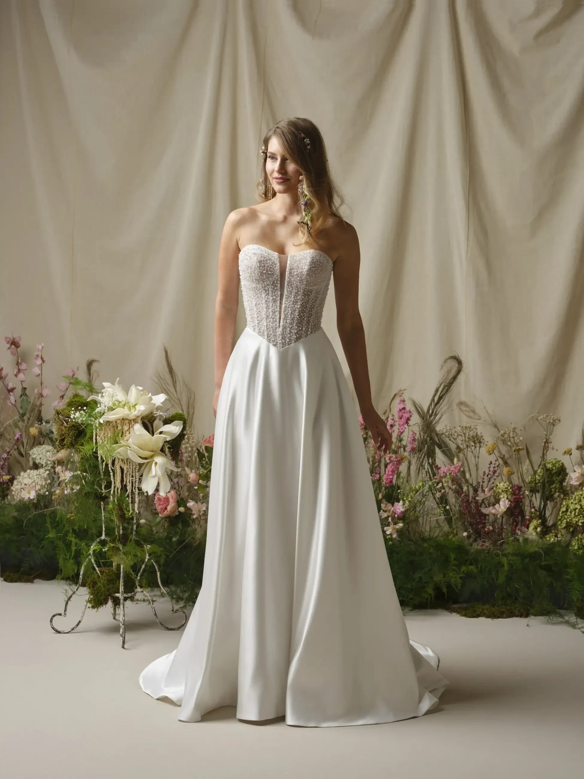 A woman in a strapless, elegant white gown with a corset bodice stands in front of a floral arrangement against a soft backdrop.