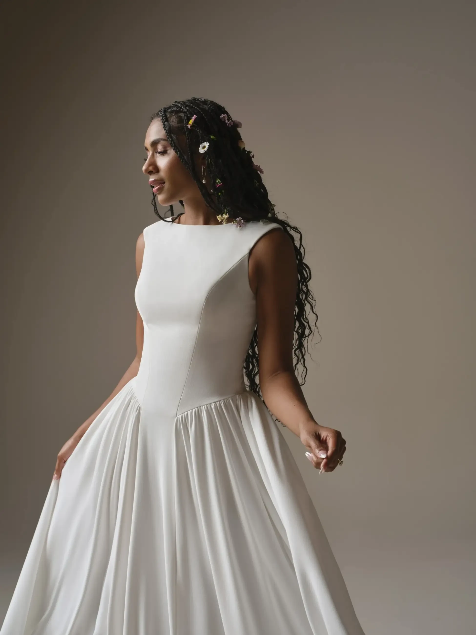 A woman in a flowing white dress with braided hair adorned with flowers stands gracefully against a simple background, exuding elegance and serenity.